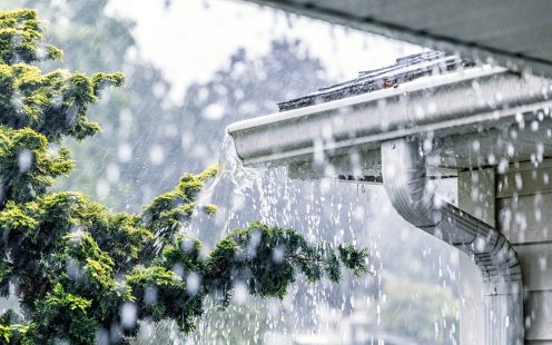 Drenching downpour rain storm water is overflowing off the tile shingle roof - streaming, rushing and splashing out over the overhanging eaves trough aluminum roof gutter system on a suburban residential colonial style house near Rochester, New York State, USA during a torrential mid-summer July downpour.